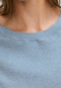 Cou du haut de la poitrine d'une personne portant un pull léger bleu clair à col rond, avec la clavicule visible et des cheveux bruns courts.