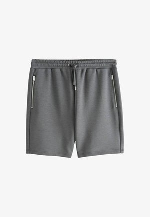 Tânăr stă cu mâinile în buzunare, purtând tricou negru, pantaloni scurți gri și adidași negri lângă o plantă mare verde în ghiveci, în fața unor pereți bej.