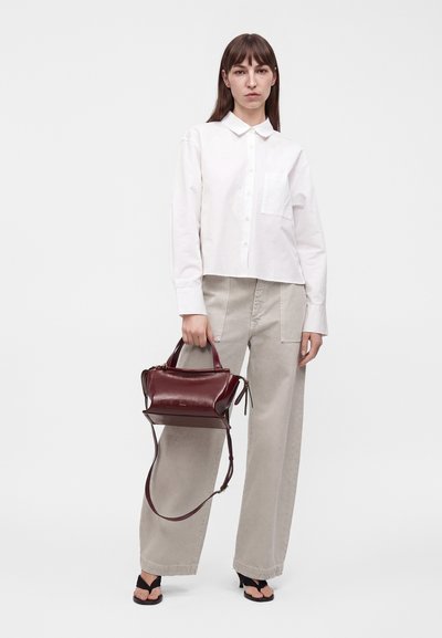 Femme en chemise blanche boutonnée et pantalon large beige, tenant un sac à main en cuir rouge foncé avec poignée supérieure et bandoulière.
