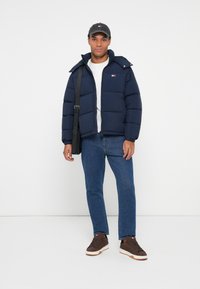 Young man wearing navy puffer jacket, blue jeans, brown sneakers, gray cap, and carrying a black shoulder bag, standing against white background.