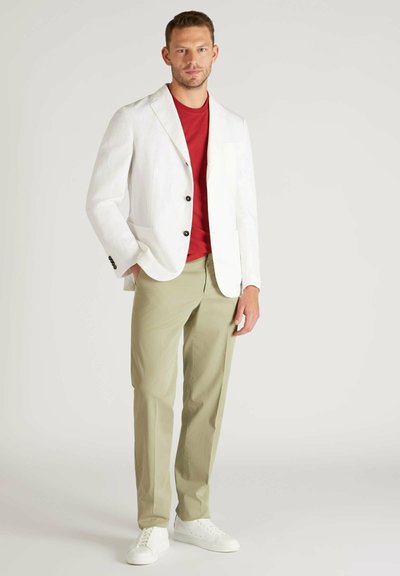 Blazer bianco in lino con bottoni neri, t-shirt rossa, pantaloni verde chiaro e sneakers bianche. Il modello si trova su uno sfondo neutro.