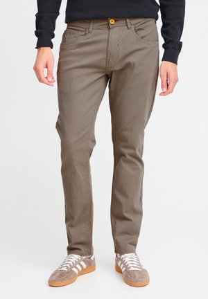 Pantalones de algodón marrón con bolsillos laterales, costuras a tono y un detalle de botón amarillo. Combinados con zapatillas de ante gris con rayas blancas.
