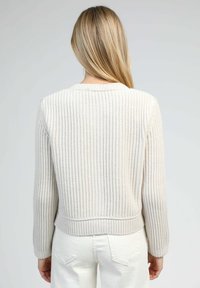 Une personne blonde portant un pull en maille côtelée crème et un pantalon blanc, vue de dos contre un fond clair uni.