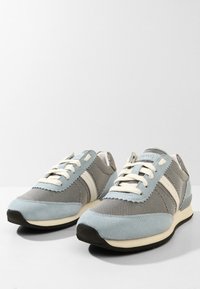 Paire de baskets grises et bleu clair avec des lacets et des rayures blanches, semelles noires, placées côte à côte sur une surface blanche.