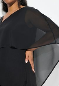 Sort bluse med en gennemsigtig overlay og lange ærmer. Stoffet fremstår glat, og der er en sideslids i kanten for ekstra detalje.