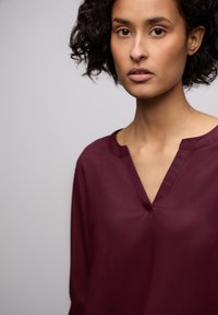 Bordeaux-bluse med V-hals, lange ærmer og glat tekstur. Har et minimalistisk design med diskrete syninger.