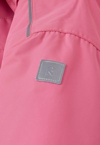 Roze stof met een textuurafwerking, voorzien van een vierkant grijs label met een reliëflogo. Stikdetails zichtbaar langs de naden.