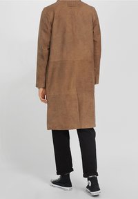 Osoba nosící dlouhý hnědý kabát, černé vyhrnuté kalhoty a černé vysoké tenisky stojí před jednoduchým pozadím.