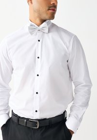 White dress shirt with a wing collar and a silver bow tie, featuring black buttons down the front. Accented by a black belt with silver buckle.