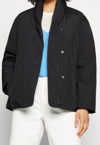 Svart vadderad jacka med hög krage, tryckknappar och frontdragkedja. Slät textur, lös passform och sidofickor synliga.