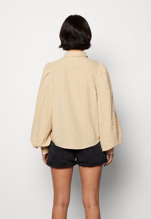 Beige sävyinen teksturoitu paita, jossa on liehukkeet pitkissä hihoissa, suora kaulus ja taakse yltävä yläosa. Pusero käytetty tummien shortsejen päällä, ja se istuu väljästi.