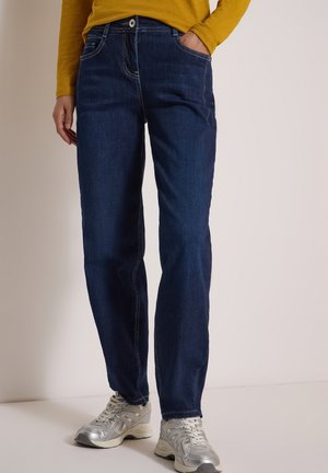 Persona che indossa jeans blu scuro a gamba dritta, sneakers argento e una maglia manica lunga color senape, con una mano in tasca, in piedi su un pavimento bianco.