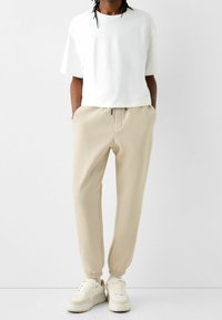 Person som bär beige joggerbyxor, vit oversized t-shirt och beige sneakers står mot en plain vit bakgrund.