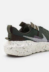 Gröna sneakers med en texturerad ovandel, elastiska remmar och en fläckig vit sula. Designen inkluderar en framträdande sidologotyp.