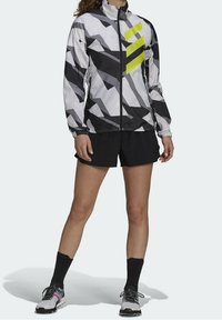 Vindjakke i sort og hvidt geometrisk mønster med lyse gule accenter. Inkluderer højtstående krave og lynlåslukning foran. Kombineret med sorte shorts.
