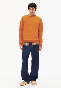 Giovane uomo con felpa arancione sopra una camicia a quadri, jeans blu scuri larghi e scarpe in stile mocassino beige, in piedi su uno sfondo bianco.