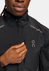 Schwarze leichte Jacke mit hohem Kragen, Reißverschluss und reflektierenden Akzenten. Verfügt über ein Logo auf der Brust und eine glatte Textur.