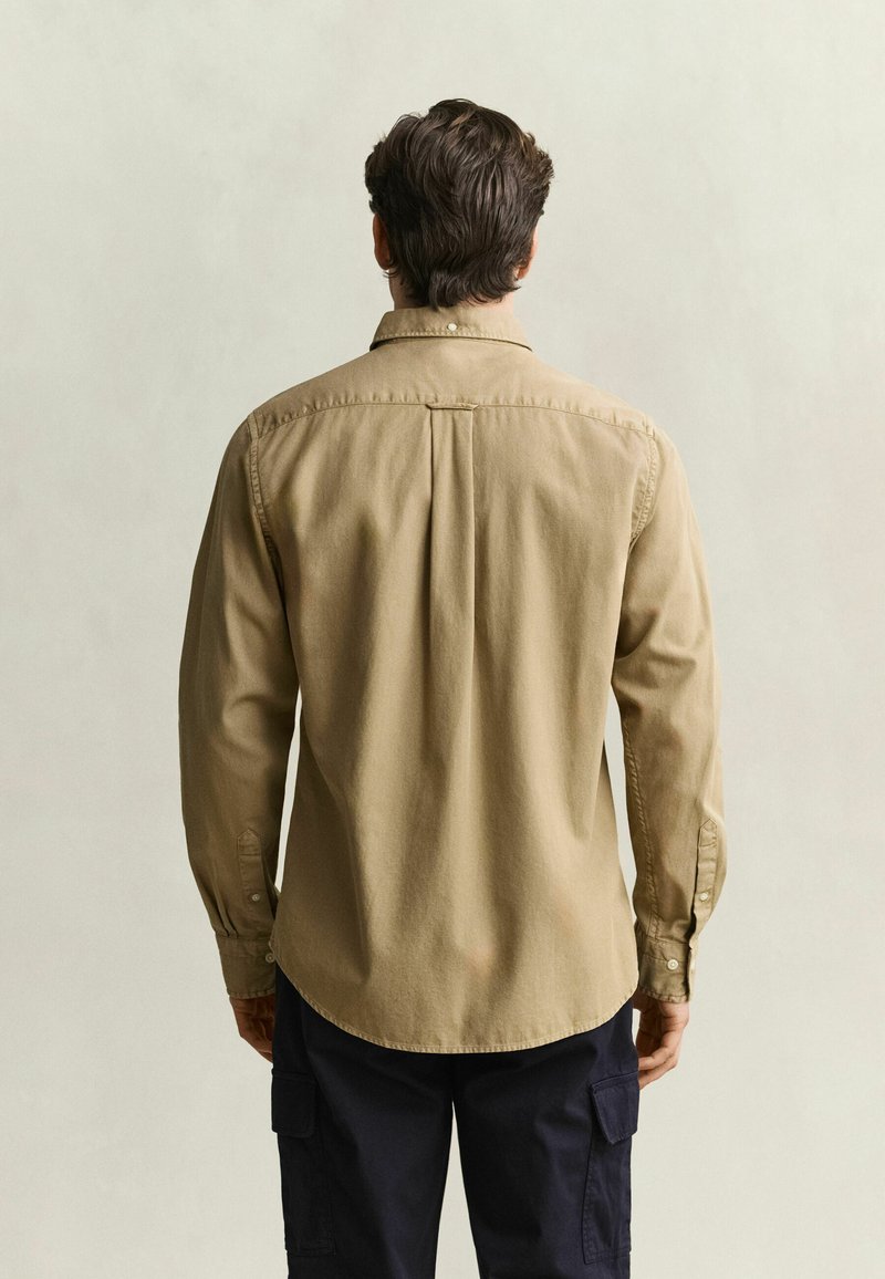 Man seen from behind wearing a beige long-sleeve shirt and dark cargo pants against a plain light background.