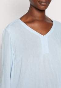 Frau trägt ein hellblaues Langarmshirt mit V-Ausschnitt, das Oberkörper und Hals vor einem schlichten Hintergrund zeigt.