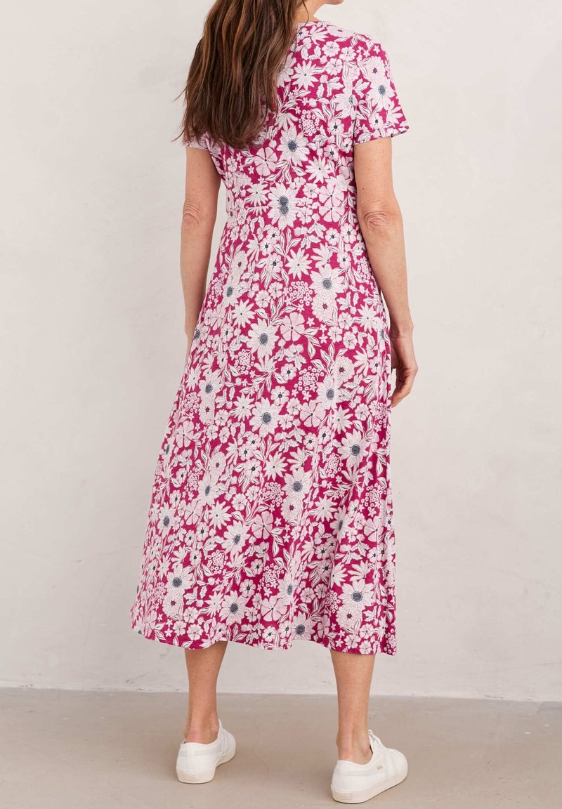 Femme portant une robe rose mi-longue à imprimé floral blanc et manches courtes, associée à des baskets blanches, debout contre un mur uni.