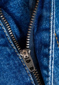 Primer plano de una cremallera metálica parcialmente abierta sobre tela de mezclilla azul con costuras amarillas visibles.