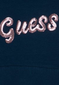 Granatowa tkanina o prążkowanej fakturze; ozdobiona błyszczącym, metalicznym różowym cekinowym logo "Guess" w stylu kursywy.