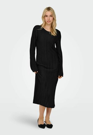 Robe noire à manches longues avec des plis verticaux, dotée d'un col rond et d'une coupe ajustée, accompagnée de chaussures noires avec des accents en nœud.