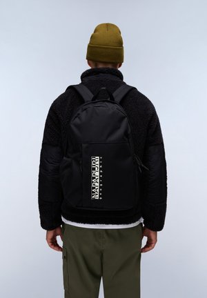 Zaino nero con una texture liscia, dotato di un logo bianco prominente, spallacci regolabili e tasche laterali in rete, indossato da una persona.