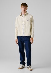 Licht beige knoppenhemd met ribbelstructuur, voorzak, gedragen over een wit T-shirt, gecombineerd met donkerblauwe spijkerbroek en sneakers.