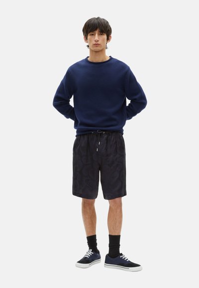 Dunkelblau gestrickter Pullover, schwarze gemusterte Shorts, schwarze Socken und Sneakers. Das Outfit zeichnet sich durch eine lockere Passform und einen Kordelzug an den Shorts aus.