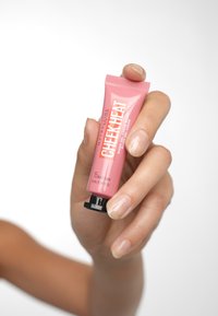 Tubetto rosa di gel blush Maybelline Cheek Heat, con tappo a vite nero e testo bianco. Mano che tiene il tubetto su uno sfondo chiaro.
