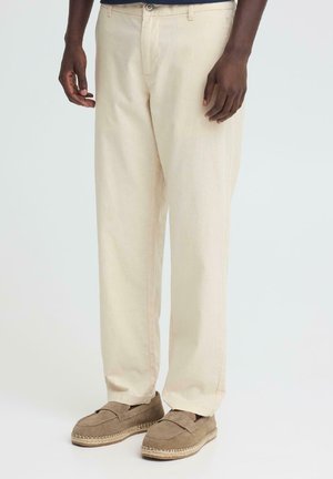 Beżowe spodnie o prostych nogawkach w połączeniu z brązowymi wsuwanymi mokasynami na stojącej osobie o ciemnym odcieniu skóry.