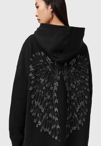 Fekete kapucnis pulóver, amelyen egy nagy, részletes ezüst színű biztonsági tű szárny grafika található a hátsó részen. Puha szövet, laza fazon és kapucni jellemzi.