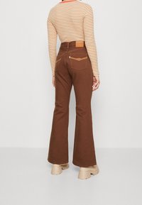 Pantalones acampanados marrones hechos de una tela con acabado texturizado; cuenta con un yugo trasero con costuras en contraste y bolsillos, combinados con zapatos de plataforma beige.