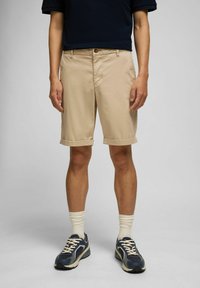 Beige Baumwollshorts mit umgeschlagenen Säumen, Fronttaschen und Knopfverschluss. Getragen mit marineblauen Turnschuhen und weißen gerippten Socken.