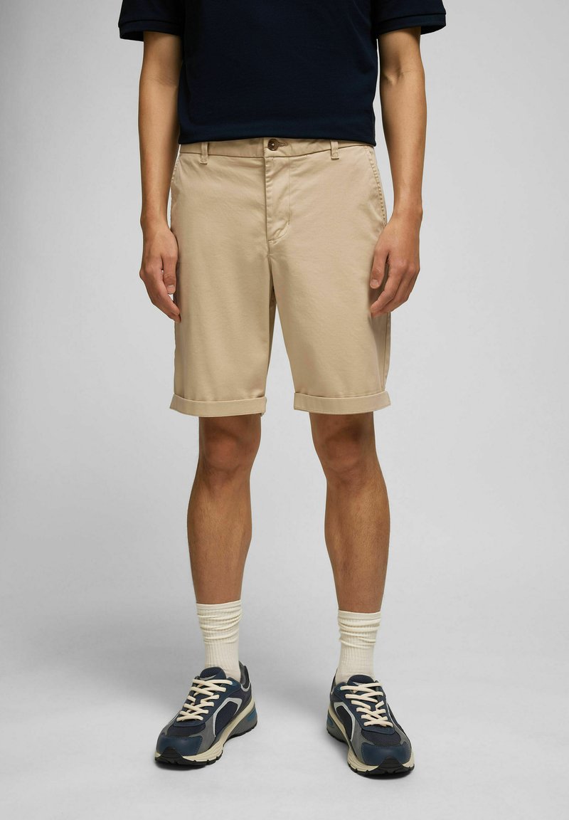 Beige Baumwollshorts mit umgeschlagenen Säumen, Fronttaschen und Knopfverschluss. Getragen mit marineblauen Turnschuhen und weißen gerippten Socken.