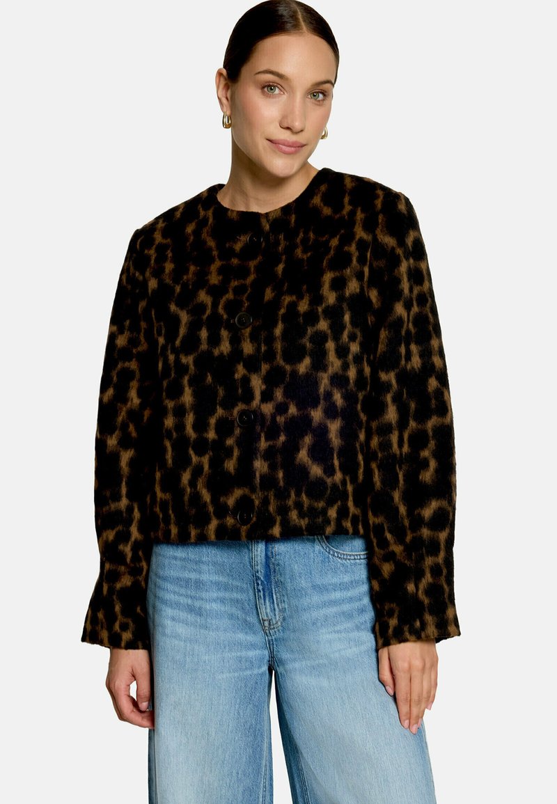 Femme portant une veste boutonnée imprimé léopard noir et marron avec un jean bleu, debout devant un fond clair uni.