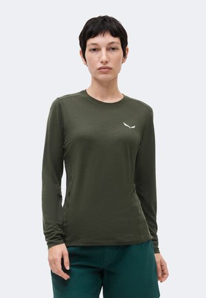 Femme aux cheveux courts et foncés portant un t-shirt à manches longues vert olive avec un petit logo blanc sur la poitrine, et un pantalon bleu sarcelle, debout devant un fond uni.