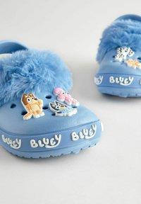 Blaue Clogs mit weichem blauen Kunstfell, ausgestattet mit bunten Charakter-Accessoires und 'BLUEY'-Branding an der Seite. Strukturierte Gummisohle.