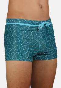 Shorts de bain teal en matériau lisse et extensible avec un motif bleu ondulé, dotés d'une taille réglable avec cordon.