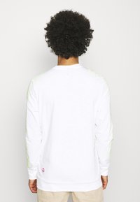 Valkoinen pitkähihainen paita, jossa on vaaleanvihreät graafiset kuviot hihoissa ja pieni vaaleanpunainen logo helmassa. Valmistettu pehmeästä, teksturoidusta materiaalista.