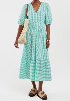 Femme portant une robe midi à carreaux verts et blancs avec des manches bouffantes, des sandales beiges à enfiler, un sac à épaule tissé et un bracelet en perles.