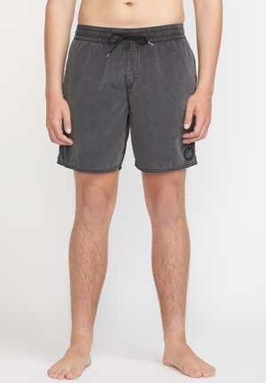 Buste et jambes d'un homme portant un short de bain gris foncé avec cordon de serrage et écusson logo sur la jambe gauche, debout pieds nus sur fond blanc.