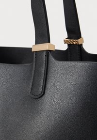 Czarna torba typu tote o fakturowanej powierzchni, wyposażona w złocone okucia przy mocowaniu uchwytu dla dodatkowego efektu.