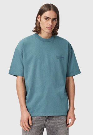 Ung mand med skulderlangt brunt hår iført en løs blågrøn t-shirt med lille mørk tekst på venstre bryst og gråvaskede jeans.