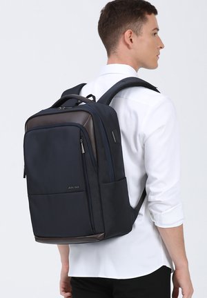 Zaino di un profondo blu navy realizzato in tessuto resistente, con dettagli in marrone, caratterizzato da più tasche, spallacci regolabili e un design elegante e rettangolare.