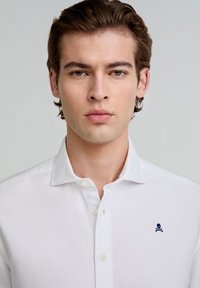 Camisa blanca de botones con cuello, que presenta un pequeño logo de calavera y huesos cruzados en la parte izquierda del pecho. Textura suave.