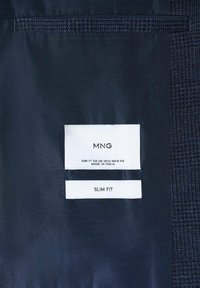 Étiquette de vêtement à l'intérieur d'une veste bleu foncé indique la marque MNG, taille EUR-IT 50 UK-USA-MEX 40, fabriqué en Inde, et style slim fit.