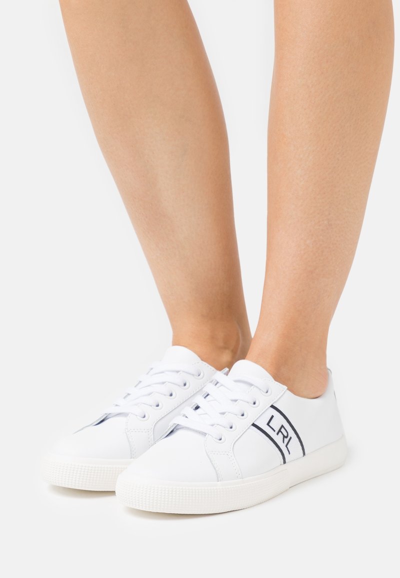 Baskets blanches à lacets avec logo "LRL" noir sur le côté, portées pieds nus sur un fond blanc uni.