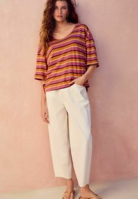 Randig, lös passform kortärmad topp i varma nyanser av orange och rosa, kombinerad med krämfärgade, vida byxor och avslappnade sandaler.